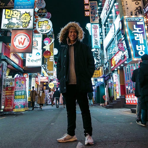 A UCR student, smiling, poses at night amid lighted signs on the streets of Hong Kong.